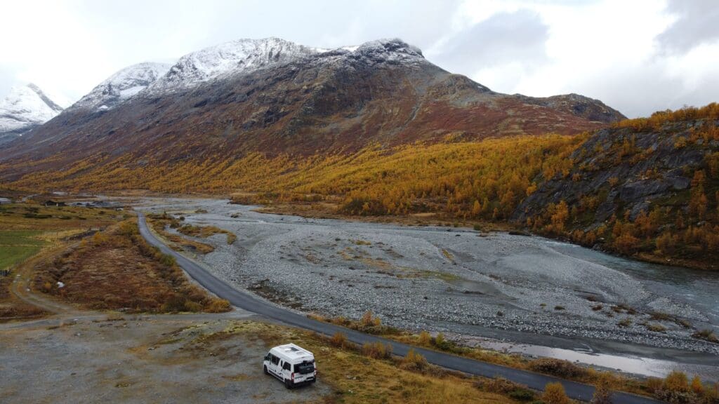 Van aménagé stationné dans une vallée norvégienne à la fin de l’automne, avec rivière pierreuse, route sinueuse, montagnes partiellement enneigées et forêts aux couleurs dorées.