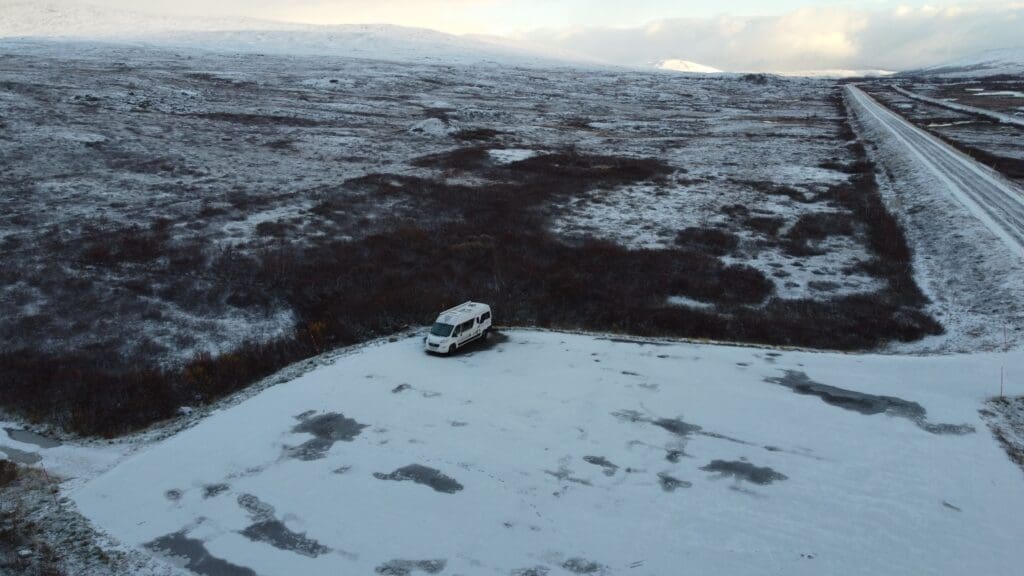 Van aménagé stationné sur une aire enneigée au cercle polaire arctique en Norvège, entouré de toundra gelée et d’une route rectiligne traversant le paysage, vue aérienne par drone.