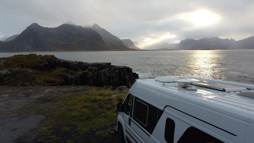 Van aménagé stationné face à la mer sur l’île de Senja en Norvège, avec montagnes abruptes en arrière-plan et lumière diffuse traversant les nuages.