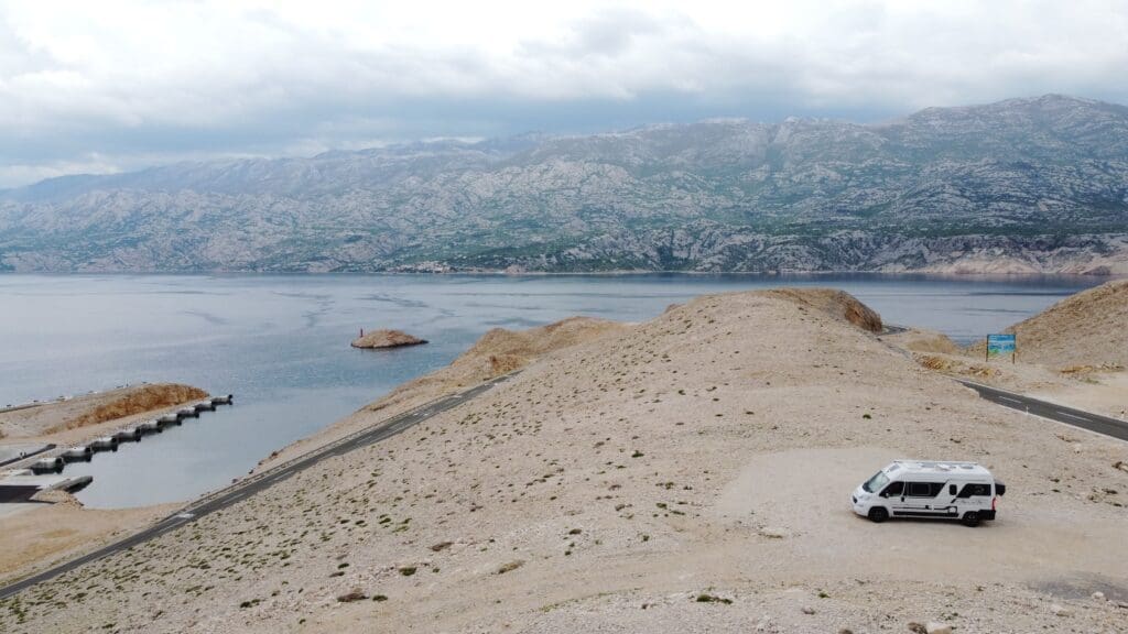 Van stationné sur un plateau rocheux aride sur l’île de Pag en Croatie, avec route sinueuse et mer calme en arrière-plan, vue aérienne par drone.