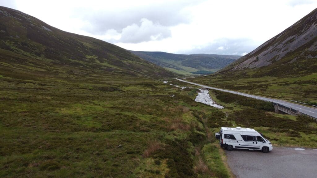 Van aménagé blanc stationné dans les Highlands écossais, au cœur d’une vallée verdoyante traversée par une rivière, entourée de collines sous un ciel nuageux.