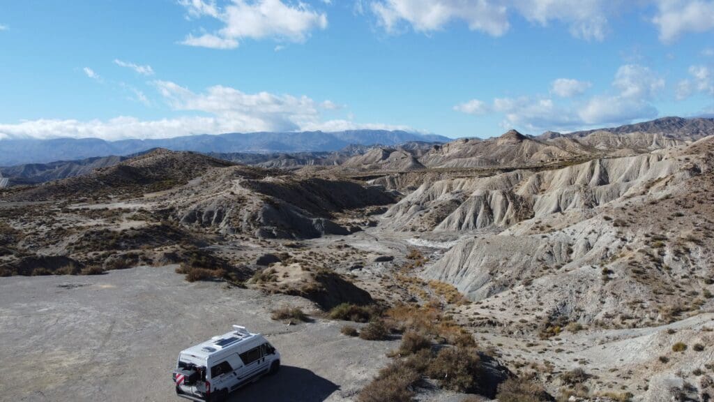 Van aménagé blanc stationné dans le désert des Bardenas Reales en Espagne, entouré de formations rocheuses arides et de paysages semi-désertiques sous un ciel bleu lumineux.