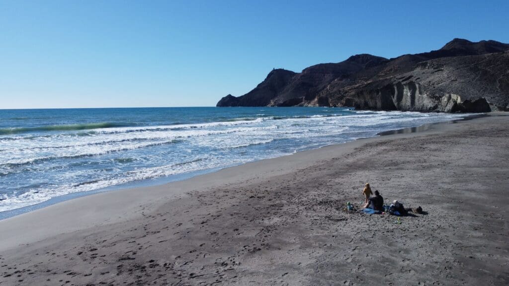 Famille installée sur une plage sauvage en Espagne, avec enfants jouant dans le sable face à la mer et des falaises rocheuses sous un ciel bleu dégagé.