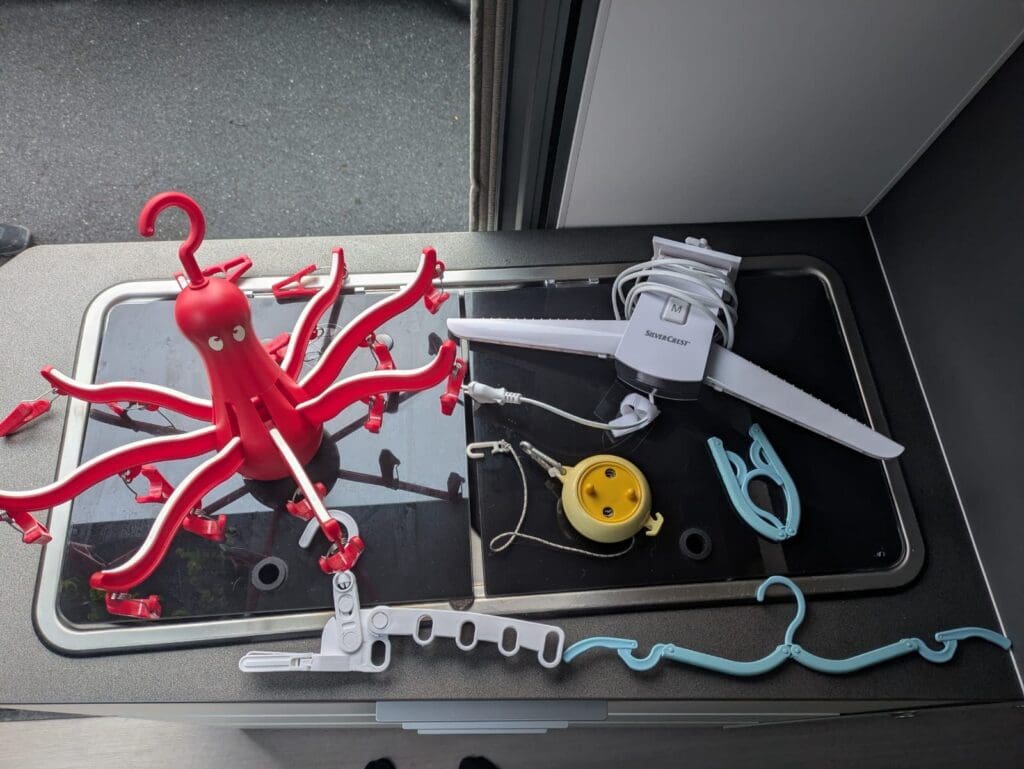 Matériel pour étendre le linge en van posé sur l’évier : étendoir “poulpe” rouge à pinces, corde à linge enroulable, mini pinces et petit appareil électrique pliant.