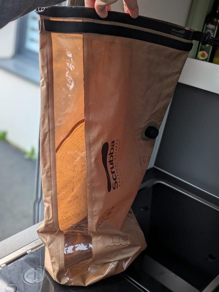 Main tenant un sac de lavage Scrubba debout dans la cuisine du van, avec une fenêtre transparente montrant l’eau à l’intérieur et une valve noire pour retirer l’air.