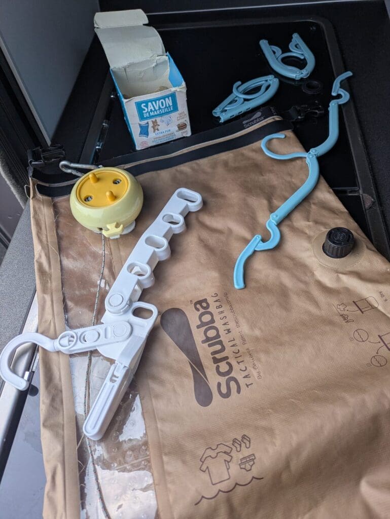 Sac de lavage Scrubba posé sur l’évier du van avec une corde à linge enroulable, un savon de Marseille, des cintres pliables bleus et un cintre multi-encoches blanc.