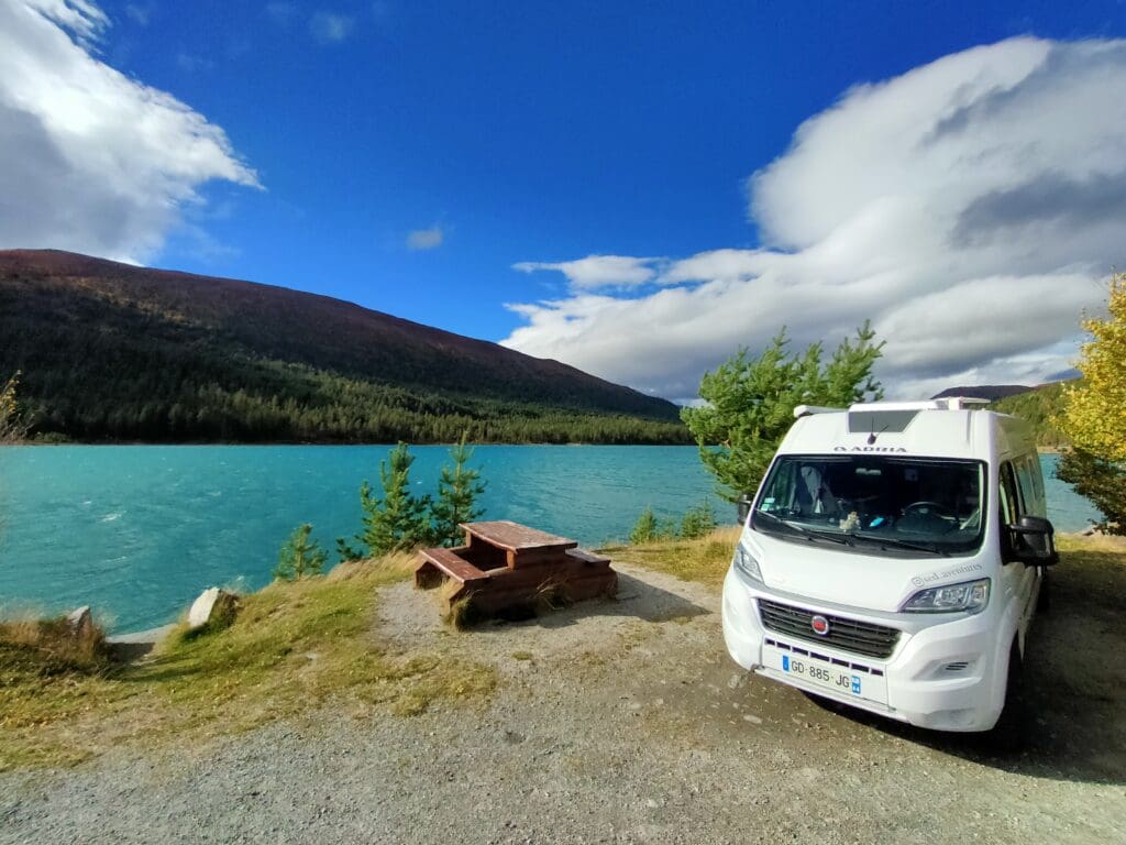 Van aménagé stationné au bord d’un lac turquoise en Norvège, entouré de montagnes et de forêt, lors d’un road trip en van.