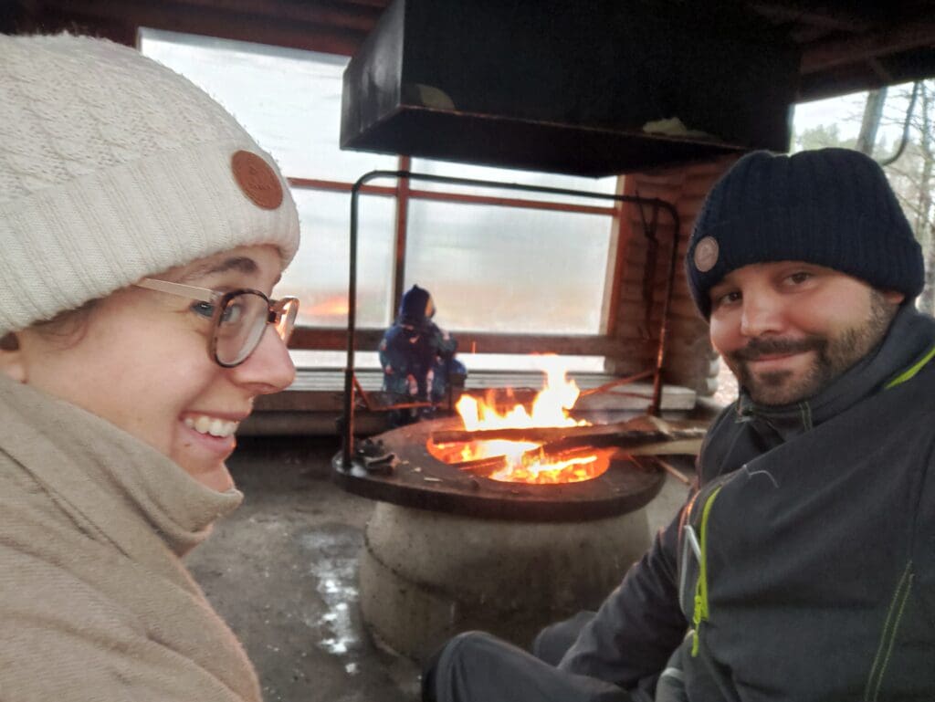 Deux adultes souriant près d’un feu de camp abrité, au bord d’un lac en Finlande en hiver, pendant un voyage en famille par temps froid.