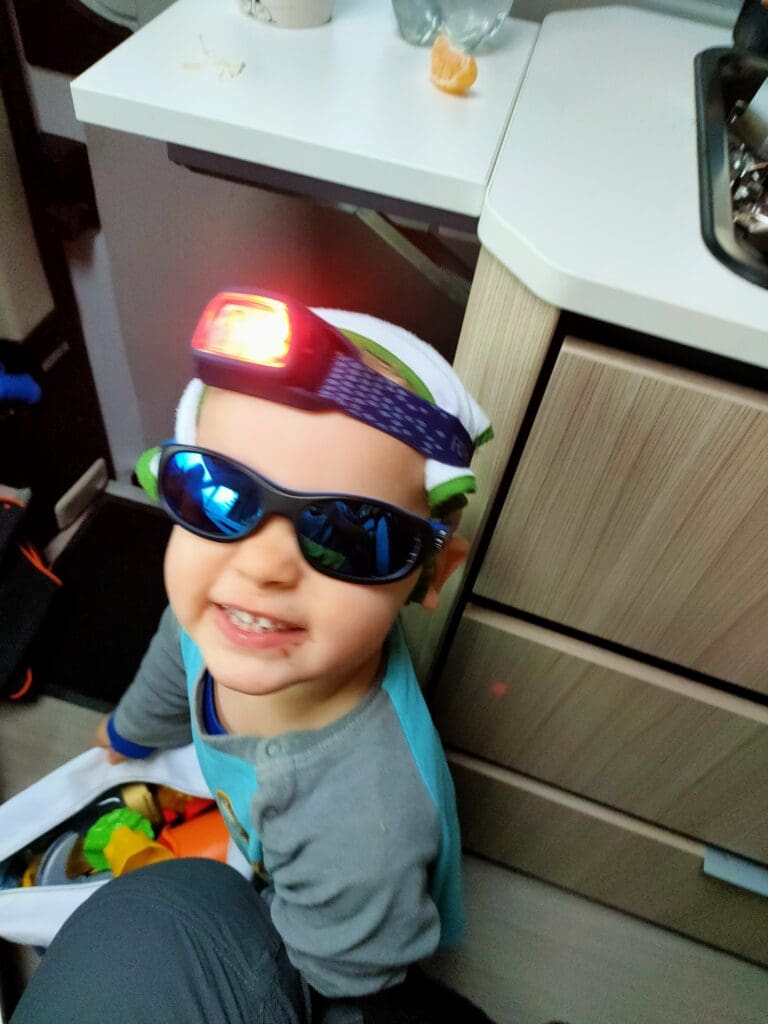 Jeune enfant souriant à l’intérieur d’un van aménagé, portant une lampe frontale et des lunettes de soleil pendant un moment de jeu.