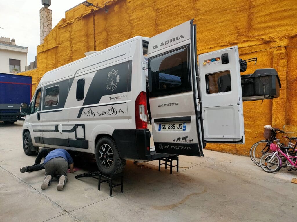 Van aménagé stationné en Espagne avec une personne réparant le chauffe-eau sous le véhicule, illustrant une panne technique en voyage en van.