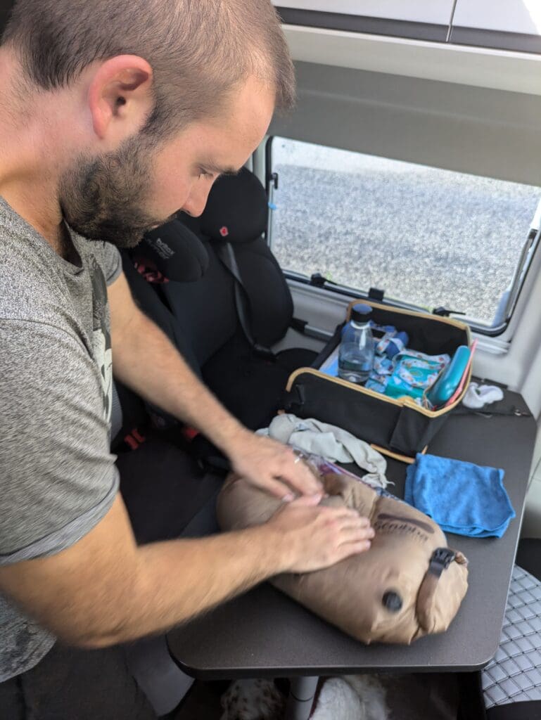 Homme debout dans un van, lavant du linge avec un sac de lavage Scrubba posé sur la table, avec une serviette et des vêtements à côté.