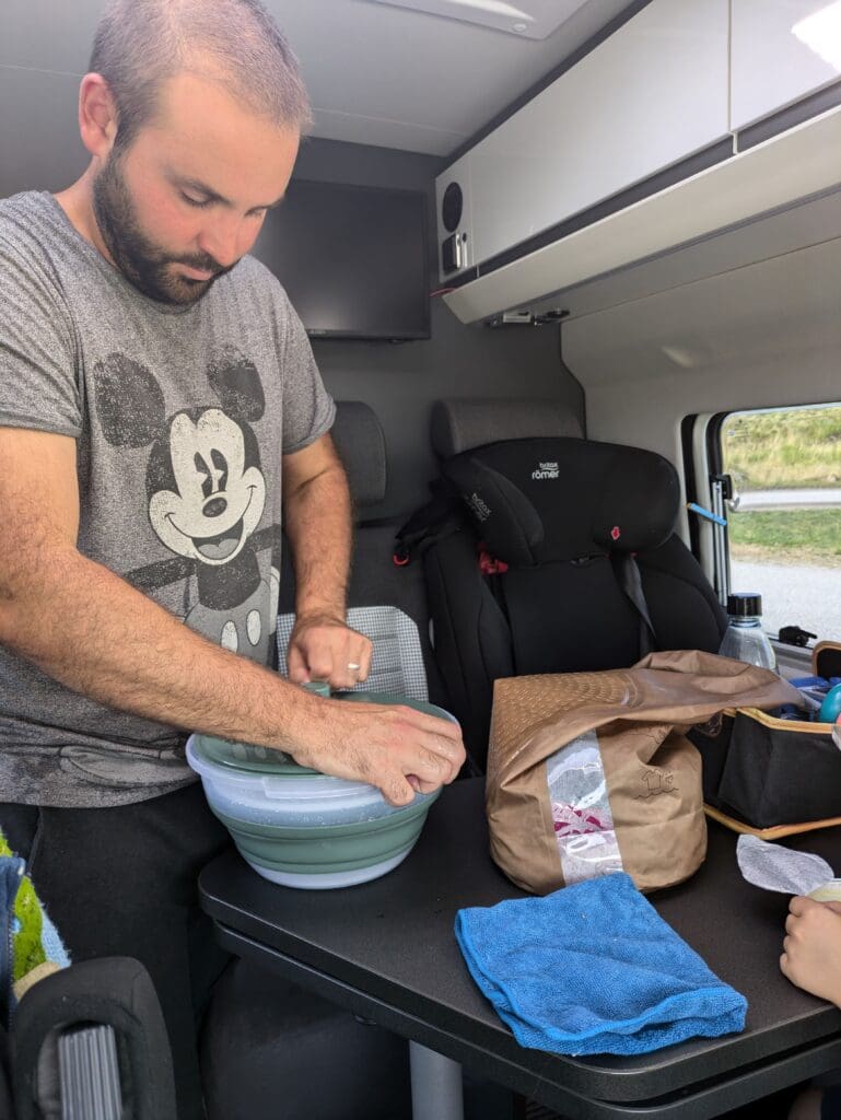 Homme dans un van utilisant une essoreuse à salade sur la table pour essorer du linge, avec un sac de lavage Scrubba et une serviette posés à côté.