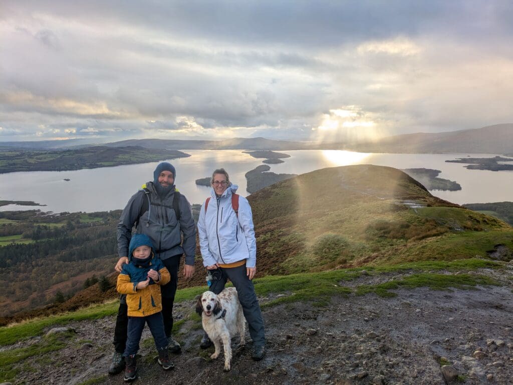 Famille de trois avec un chien au sommet d’une colline, vue sur un grand lac et des îles sous un ciel nuageux avec des rayons de soleil.