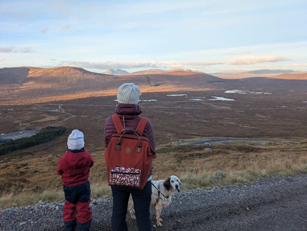 Adulte et enfant vus de dos sur un point de vue en montagne, avec un chien en laisse, face à une vaste vallée parsemée de petits lacs sous un ciel clair.