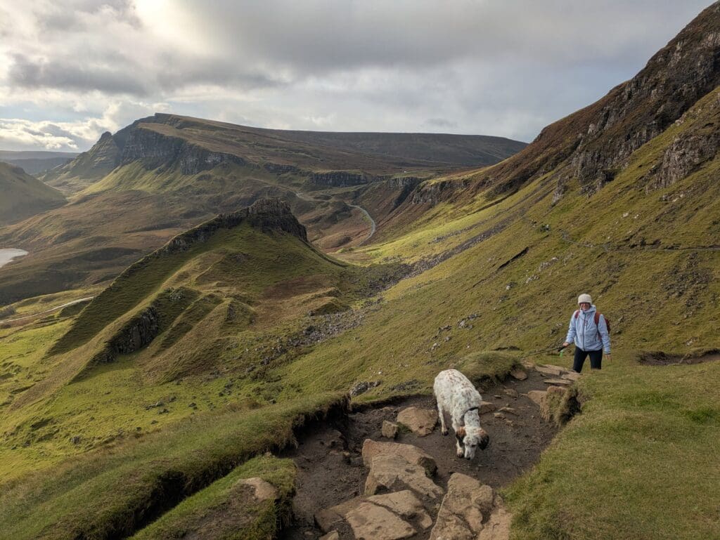 Chien blanc tacheté sur un sentier boueux avec des pierres, tandis qu’une personne marche derrière, dans une vallée de montagnes verdoyantes sous un ciel nuageux.