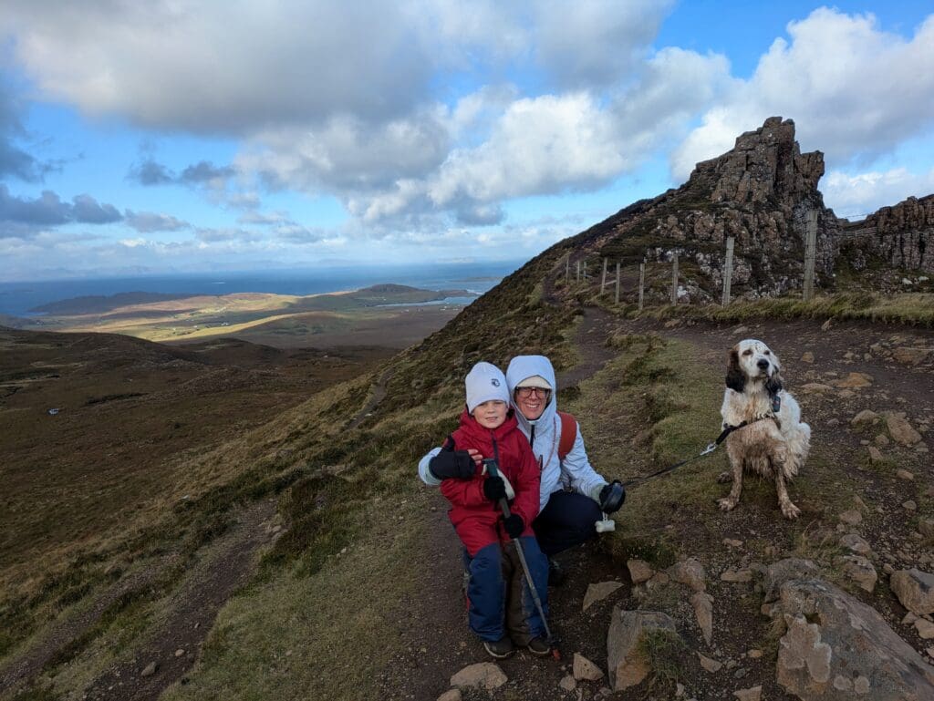 Adulte et enfant accroupis sur un sentier de montagne avec un chien en laisse, près d’un rocher, vue sur la mer et des collines sous un ciel nuageux.