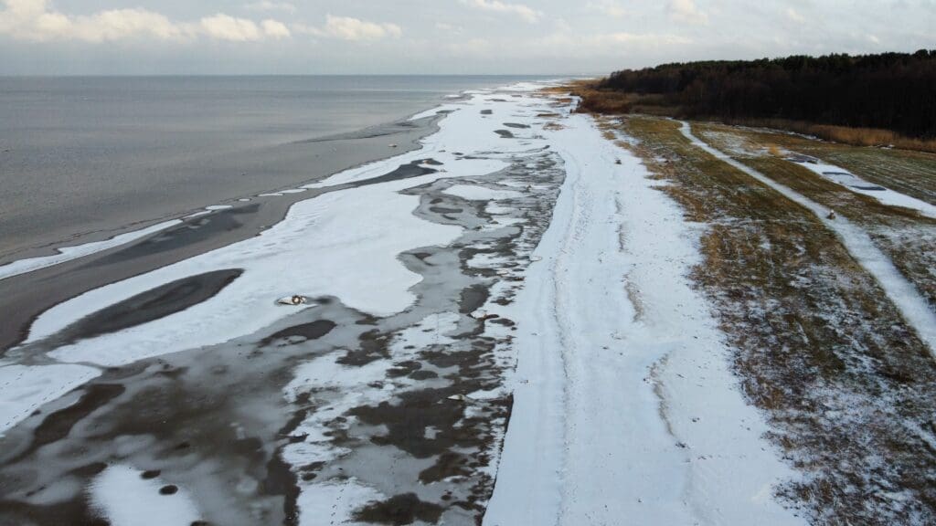 Plage en Estonie partiellement recouverte de neige, avec rivage gelé, mer calme et bande forestière en arrière-plan, vue aérienne par drone.