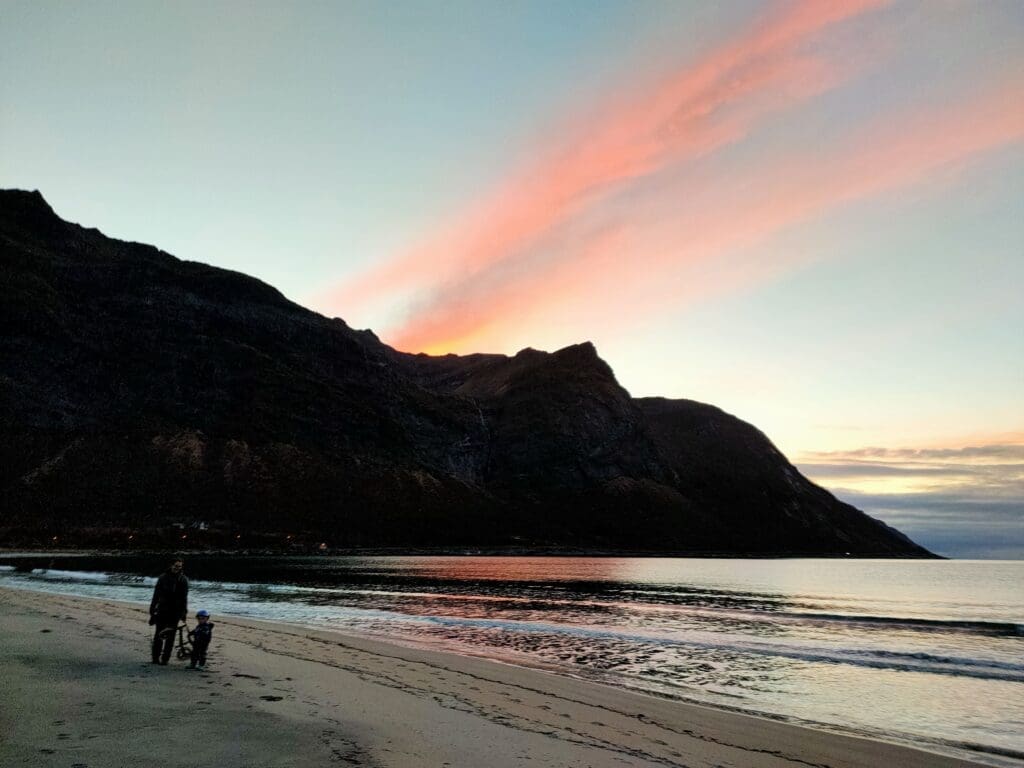 Parent et enfant marchant sur la plage de Senja en Norvège au coucher du soleil, ciel rose et orange sur les montagnes arctiques, road trip en famille