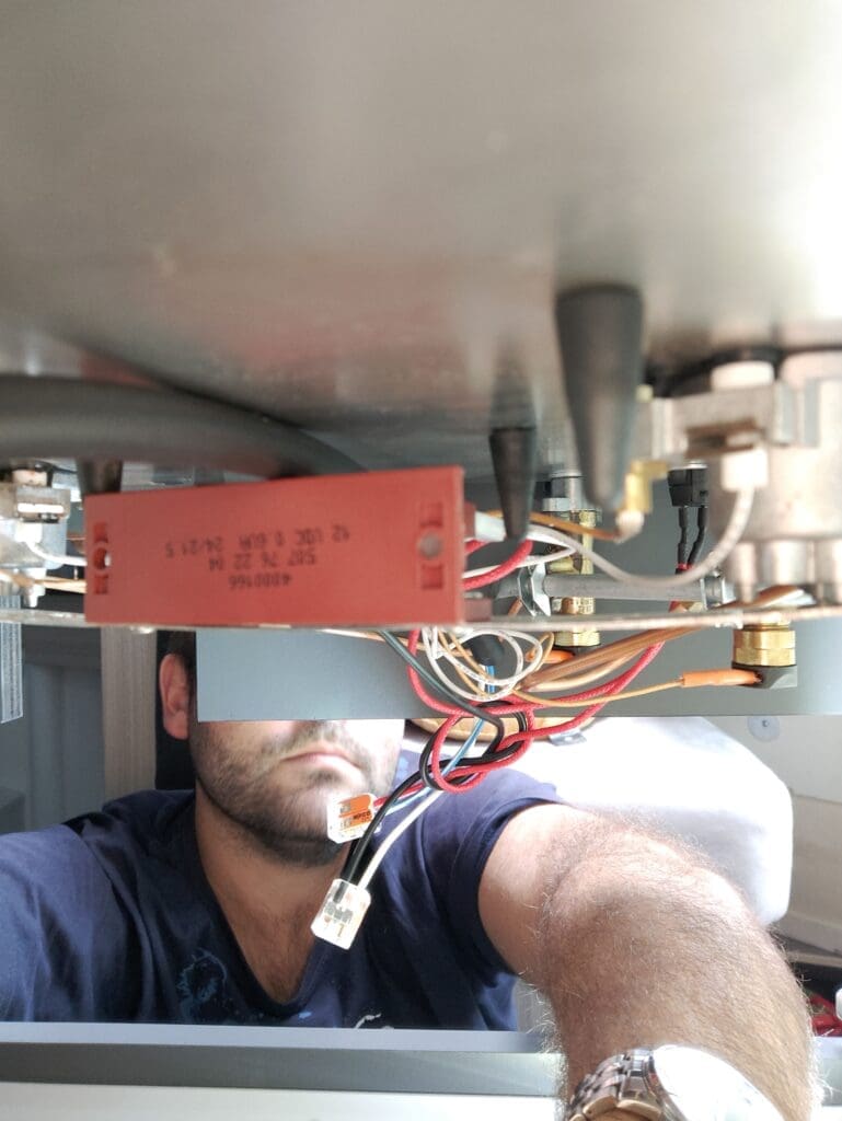 Kakou penché sous les plaques de cuisson du van pour réparer la panne d'allume-gaz, câbles et composants visibles — mécanique vanlife