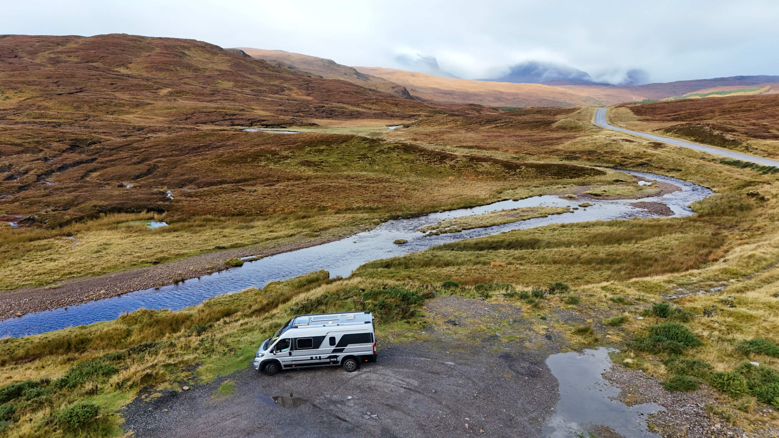 Vue aérienne d'un van aménagé près d'une rivière sinueuse, au milieu des landes et montagnes d'Écosse sous un ciel nuageux.