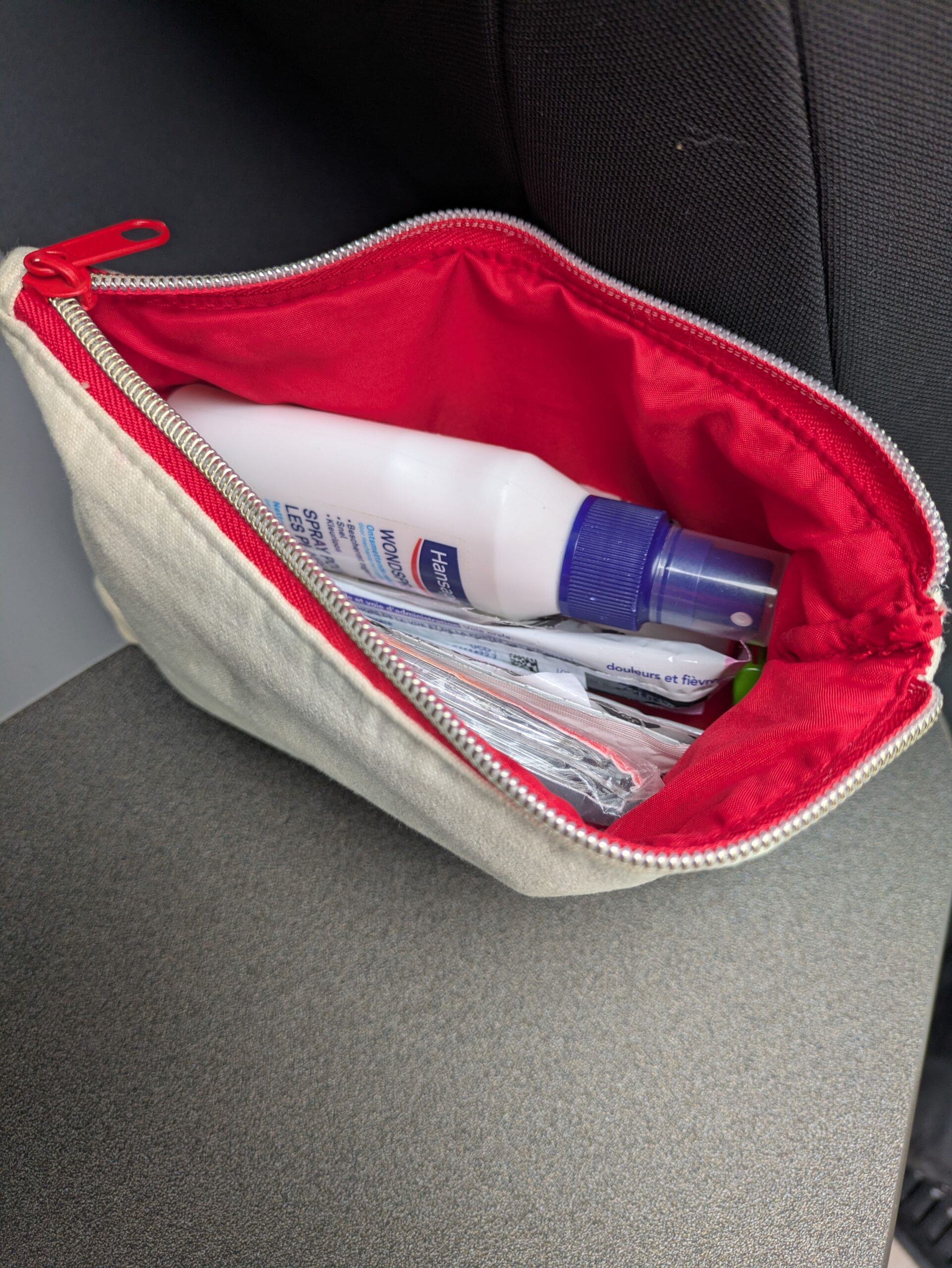 Petite pochette beige à fermeture éclair, doublée de rouge, contenant un spray et plusieurs produits de premiers secours.