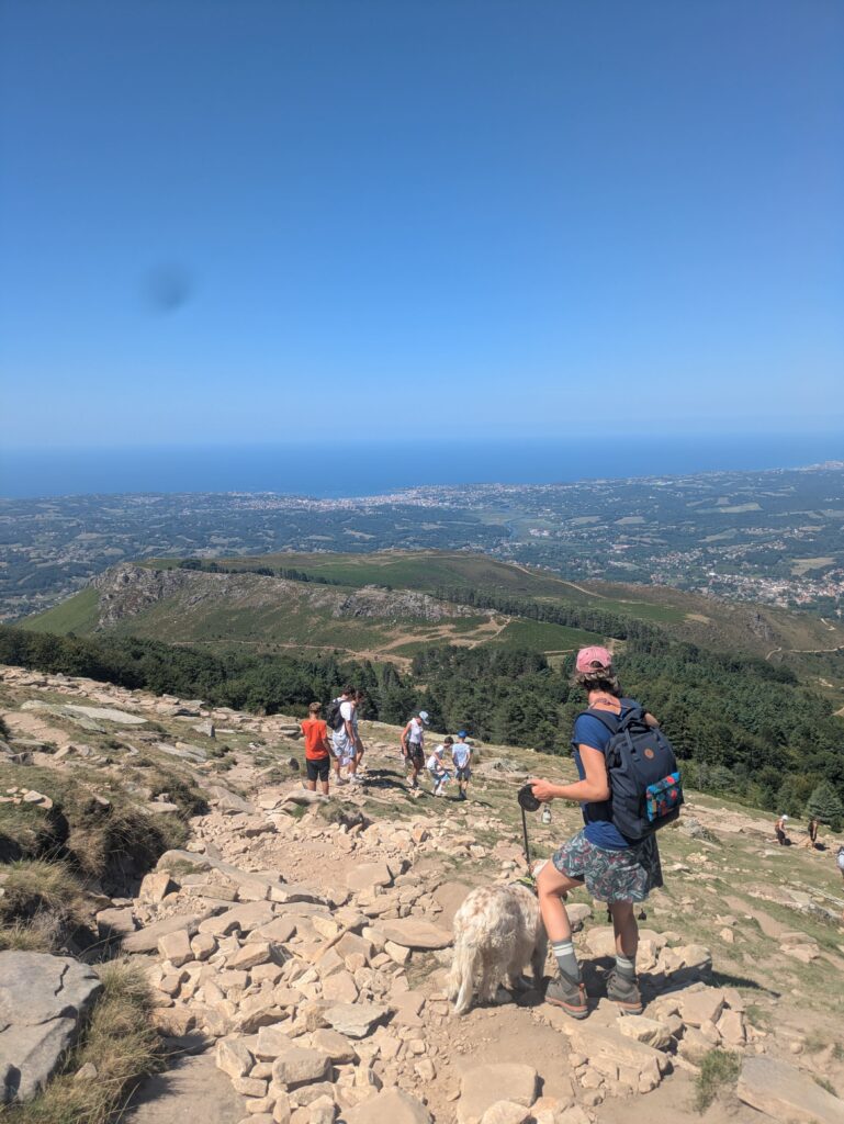 Randonneuse accompagnée d’un chien blanc sur un sentier rocheux de la Rhune, dans le Pays basque, avec une vue sur la côte atlantique.