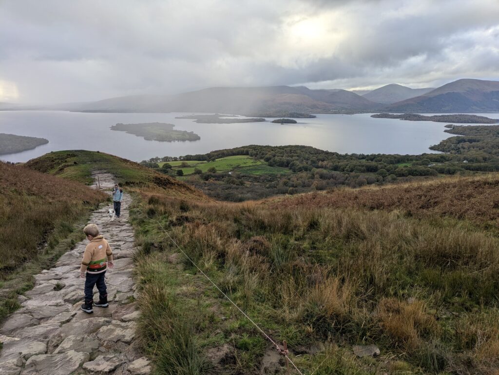 Enfant montant un sentier de pierres à Conic Hill, avec une randonneuse et son chien face aux îles du Loch Lomond en Écosse.