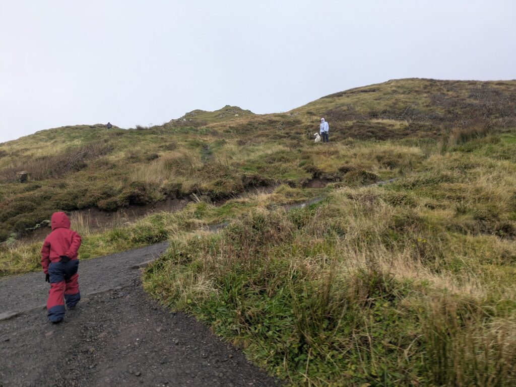 Enfant en tenue rouge marchant sur un sentier de l'Old Man of Storr, vers une randonneuse et un chien, sur l'île de Skye.