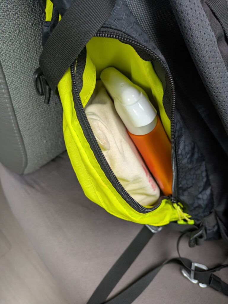 Petite trousse de secours ouverte dans une poche jaune vif d’un sac à dos gris, contenant un flacon orange et une pochette textile.