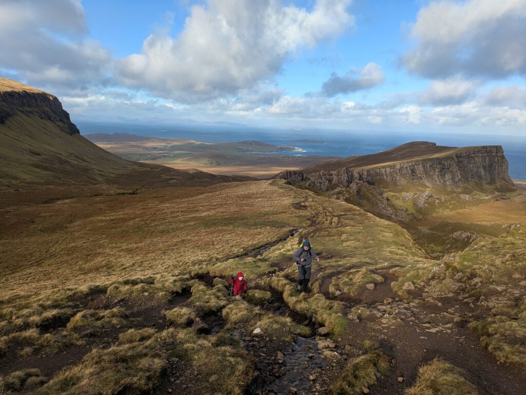 Deux randonneurs gravissent un sentier escarpé du Quiraing, avec vue sur les falaises, les vallées et la mer au nord de l'île de Skye.
