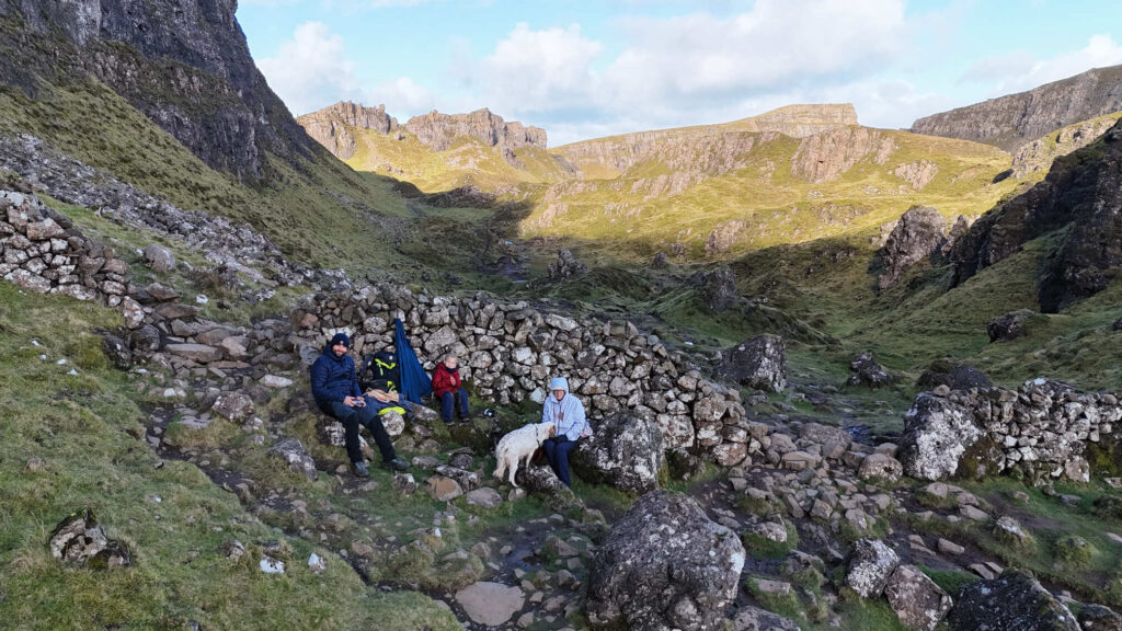 Famille et chien faisant une pause près d'un ancien muret de pierres au Quiraing, entourés de falaises et de montagnes verdoyantes sur l'île de Skye.