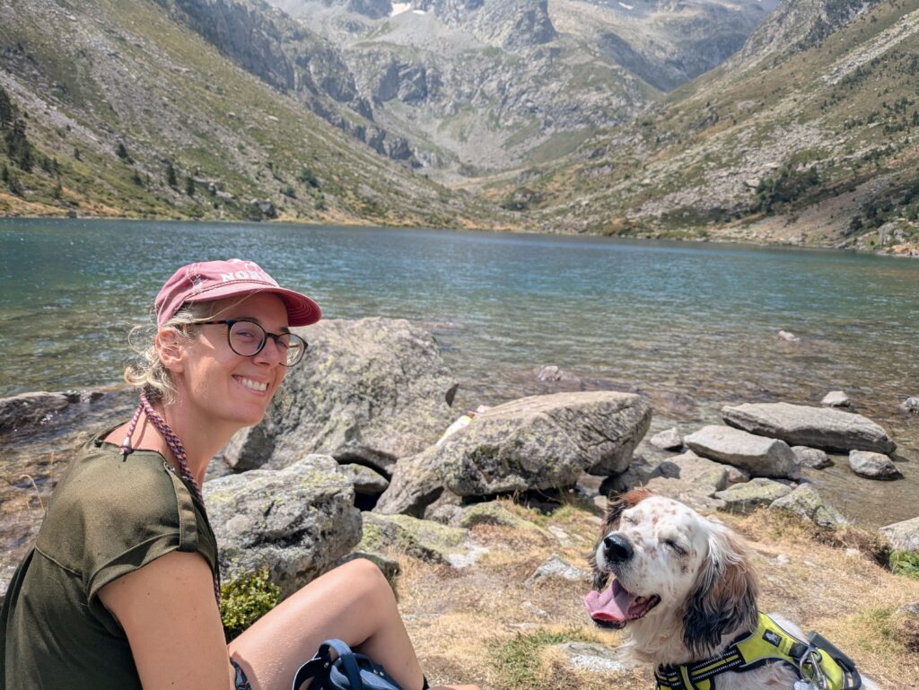 Femme souriante assise près d’un lac de montagne avec un setter anglais blanc équipé d’un harnais, devant un paysage rocheux.