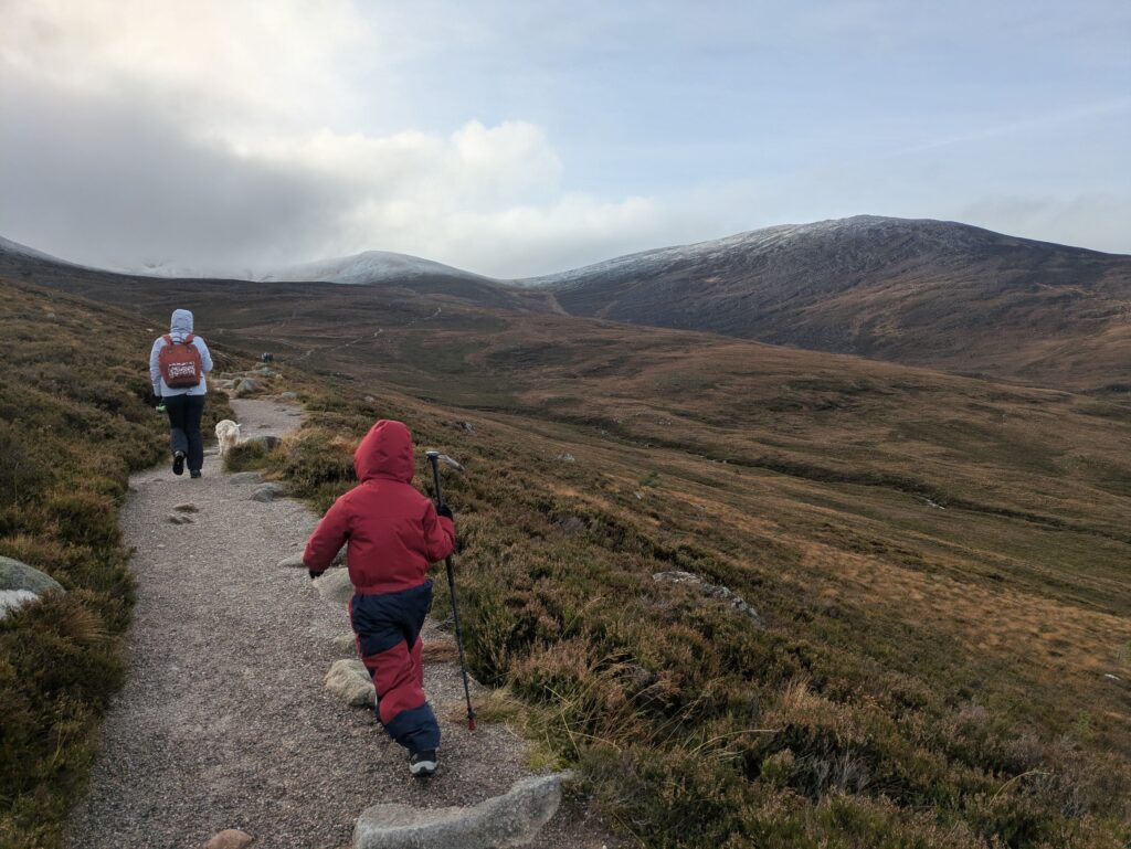 Enfant en combinaison rouge suivant une randonneuse et un chien sur un sentier des Cairngorms, face aux montagnes légèrement enneigées d'Écosse.