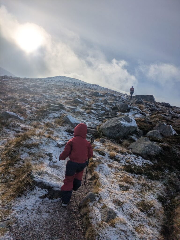 Enfant équipé d'un bâton de randonnée gravissant un sentier rocheux légèrement enneigé dans les montagnes des Cairngorms, en Écosse.