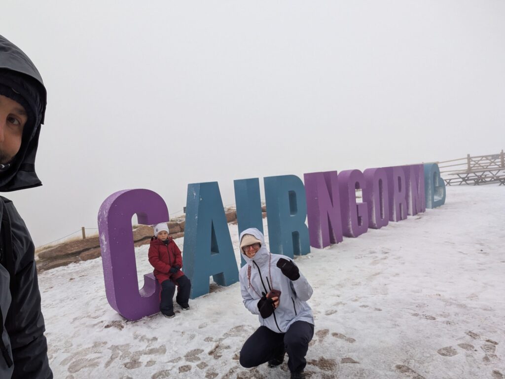 Famille photographiée près des grandes lettres colorées CAIRNGORM, sur un sol enneigé et dans la brume, à Cairngorm Mountain en Écosse.